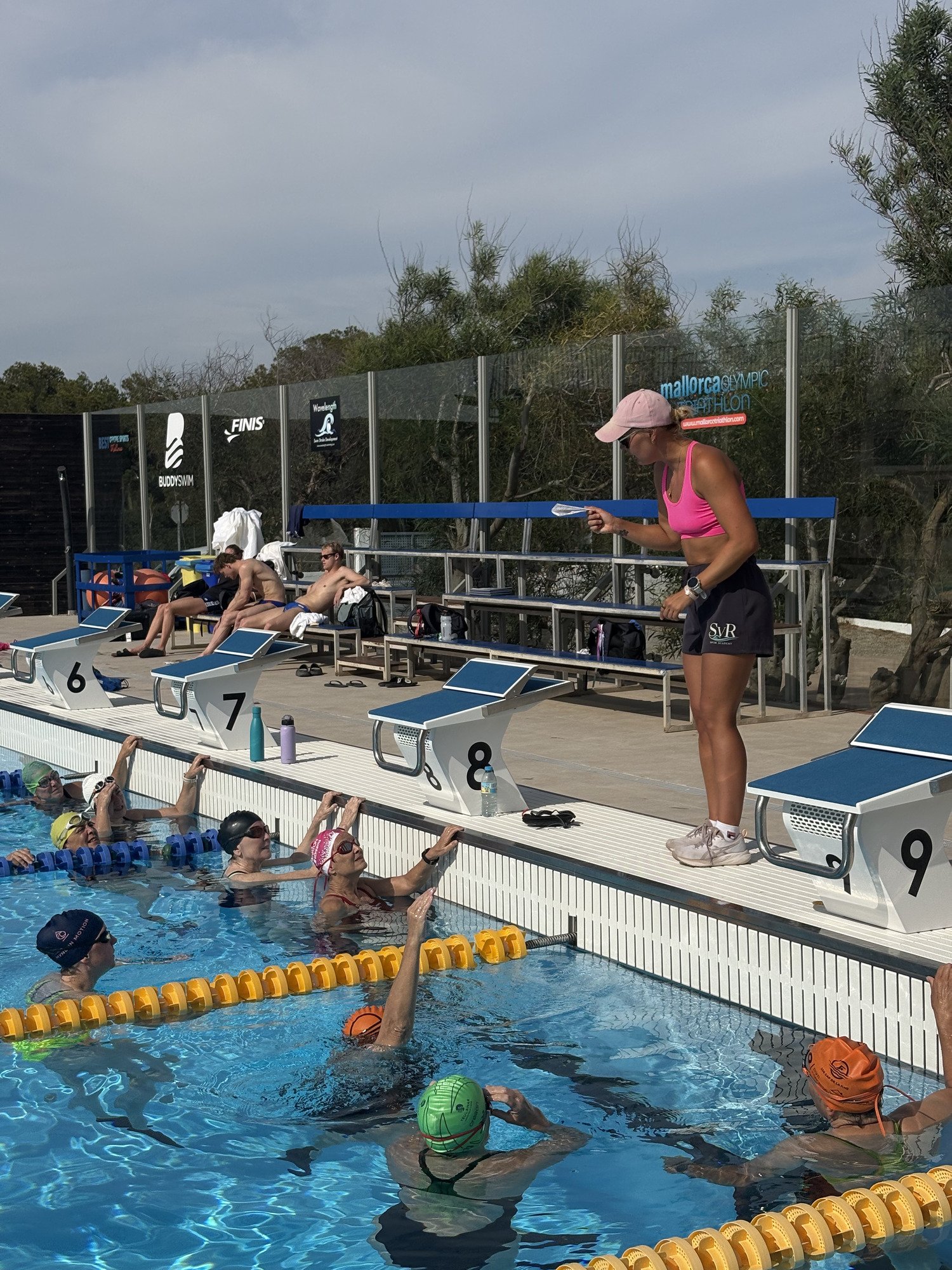Sharon coaching swimmers at the outdoor pool in Mallorca, wearing a pink top.