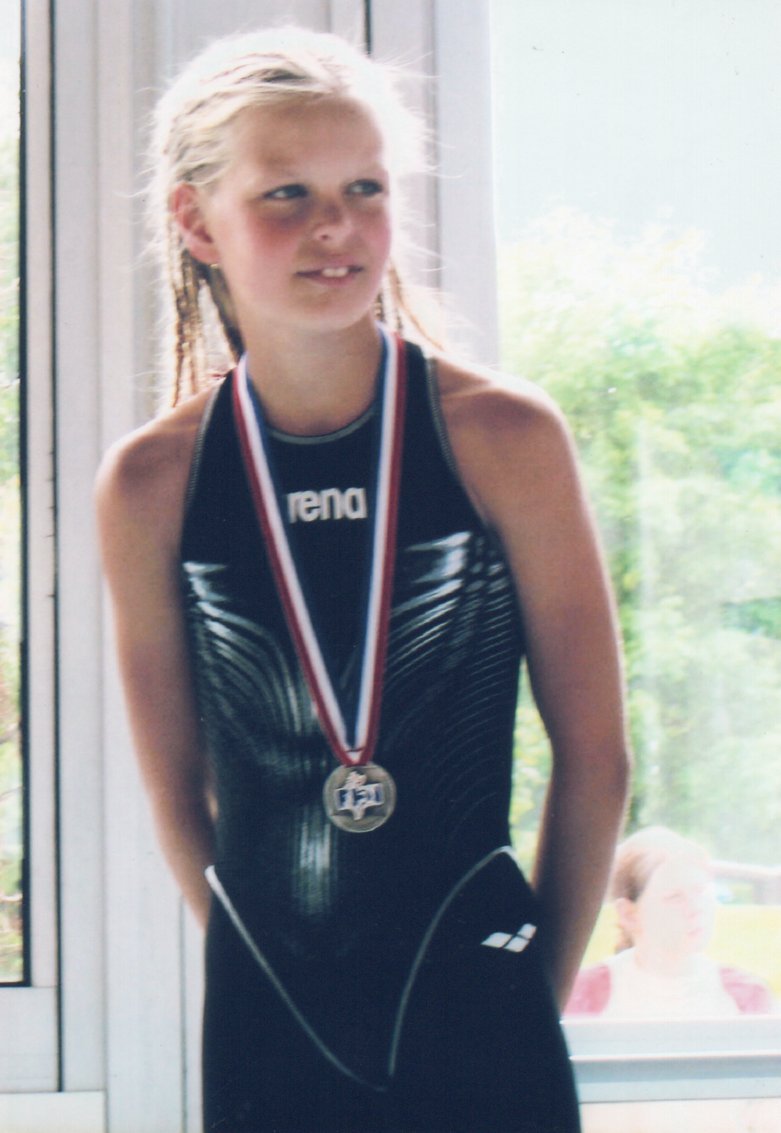 A young swimmer wearing a medal and holding a swim cap beside a window.